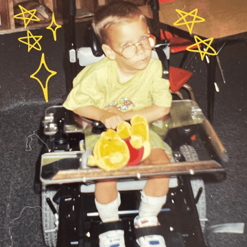 Young David Stoner wearing a yellow shirt and shorts and a pair of glasses in a power wheelchair holding a winnie the pooh teddy bear