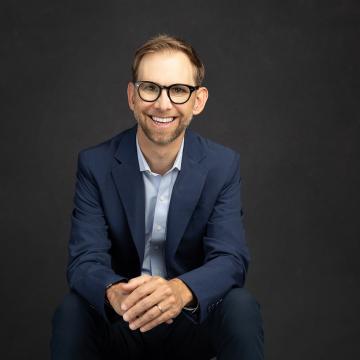 Michael Kutcher seated wearing a dark blazer with light shirt and dark rimmed glasses