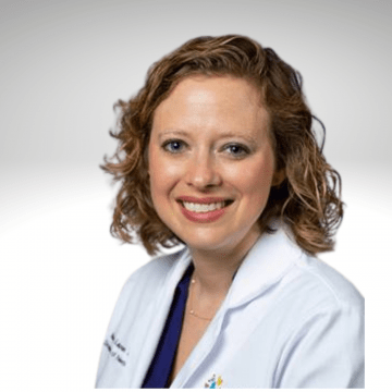 Mary Lauren has curly blond shoulder length hair and is wearing a white lab coat. She is smiling