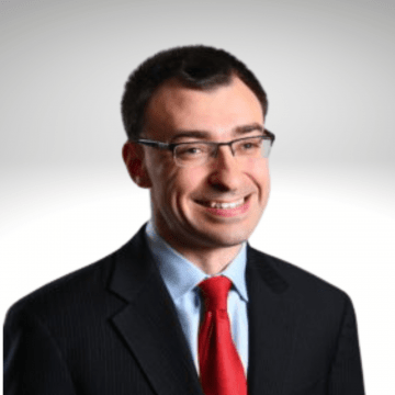 Jason has dark brown hair and is smiling wearing a blue shirt, red tie and a blazer.