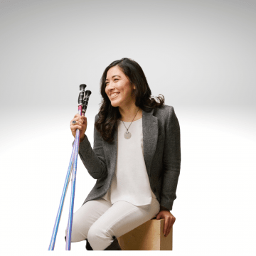 Xian Horn is sitting on a bench smiling, wearing white pants, white shirt and a brown blazer. She has walking sticks in her right hand. She has long brown hair and is smiling.