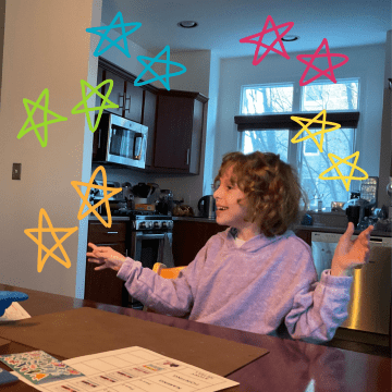 A young girl with medium length curly brown hair is smiling with her arms outstretched while wearing a lilac shirt at a kitchen table in a kitchen of a home.