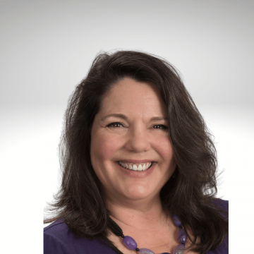 Mary Gannotti has long brown hair and is smiling. She is wear a purple top and a purple beaded necklace.