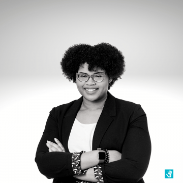 Minelli Vasques has curly brown hair, she is smiling and wearing glasses. She has a white shirt and a black blazer with her arms crossed in front of her. 