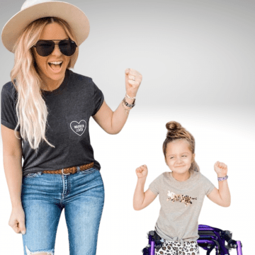 Fifi is a little girl standing in front of her purple walker wearing a grey t-shirt with her hands fisted in the air. She is smiling and has brown hair. She is next to her mom who is wearing a white cowboy hat, dark glasses, has long blonde hair and is smiling wearing a grey shirt and jeans. 