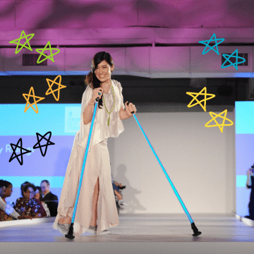 A woman with long brown hair wearing a white flowing dress is on a fashion runway using bright blue poles to walk