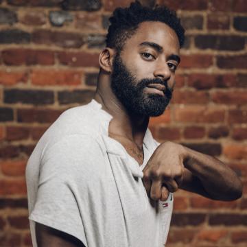 Jerron is standing in front of a brick wall wearing a white shirt with his left hand and arm up in front of his chest.