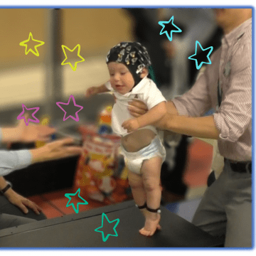 A baby wearing an eeg cap is being held up by a man while he tries to take a step towards a woman with her arms outstretched