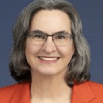 Dr. Kripke has shoulder length salt and pepper hair and is smiling wearing glasses and an orange blazer
