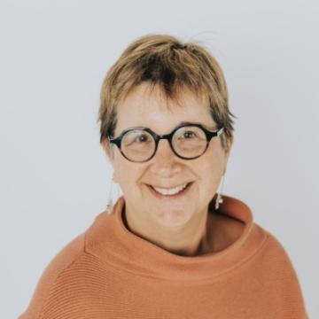 Ginny Paleg headshot. She has short brown hair and is wearing glasses and an orange turtleneck.
