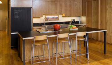 Photo of modern kitchen with wooden paneling. Island bench with 3 stools and black bench top