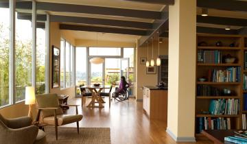 A photo of a modern home with open plan living looking from the lounge room to the dining area and kitchen. There is a young women in a manual wheelchair in the dining room.