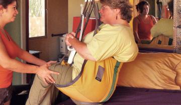 A woman using an overhead hoist with tracking system to transfer from bed to wheelchair with assistance.
