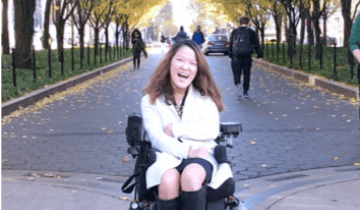 Writer Sarah Kim sitting in her power wheelchair in front of an avenue of trees