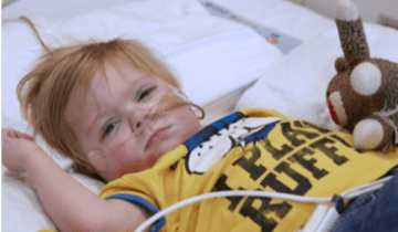 Young child lyign in hospital bed wearing yellow t-shirt and holding a stuffed toy