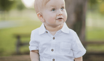 Owen (Age 2) sitting outside under a tree in a white shirt and brown shorts looking up and smiling into the distance