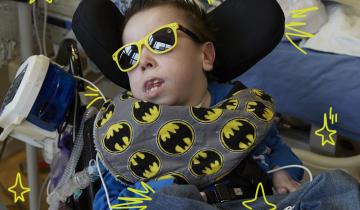 Photo of a young man in a blue shirt, wearing dark sunglasses with yellow rims reclining in a wheelchair with a batman printed neck pillow around his neck. He is in a hospital room and has a feeding tube attached to the wheelchair.