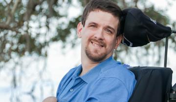 Photo of David Stoner, wearing a blue shirt, sitting in his black wheel chair with a tree in the background.