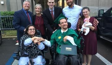Photograph of a young woman and young man wearing a graduation cap and gown in wheelchairs holding hands with five individuals standing up behind them. One individual is holding an infant.