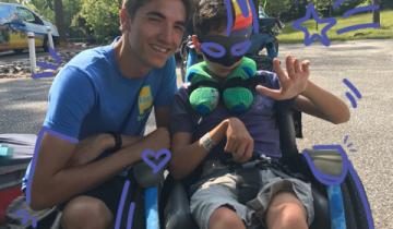 Photo of two young men in blue shirts sitting next to each other outdoors, one is in a wheelchair wearing a Mardi Gras mask and waving.
