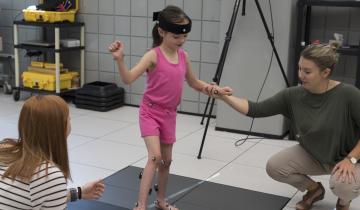 Young girl in a pink tank top and shorts holding hands with a woman in a grey shirt and tan pants while balancing on a grey mat