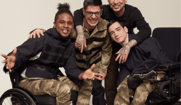 Photo of 4 men: From left to right - Black man, smiling wearing a black turleneck and camo pants seated in a wheelchair next to a white man leaning over smiling with dark hair wearing camo, next to an asian man with dark hair standing, smiling in black turtleneck, next to a white man seated wearing a black turtleneck and camo pants seated in a wheelchair.