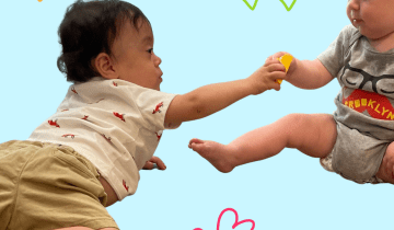 Image of a baby with dark hair and tan skin on his belly reaching to give a block to another baby sitting up with a bald head wearing a grey onesie