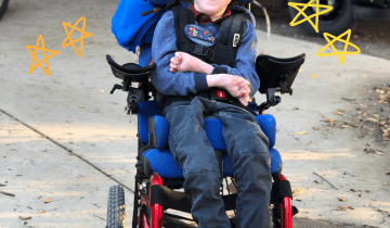 Boy with brown hair wearing a blue shirt and blue pants in a red wheelchair smiling in front of a school bus