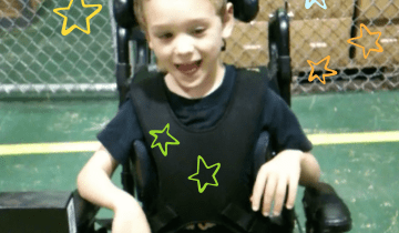 Bower in a power wheelchair wearing a black shirt and tan pants smiling and driving with a head array