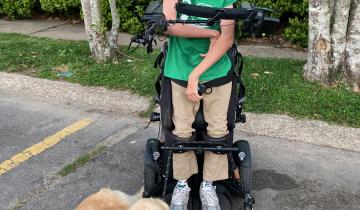Bower wearing a green shirt and tan pants standing in his wheelchair smiling with his golden retriever everest