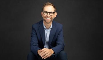 Michael Kutcher seated wearing a dark blazer with light shirt and dark rimmed glasses
