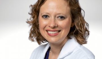 Mary Lauren has curly blond shoulder length hair and is wearing a white lab coat. She is smiling