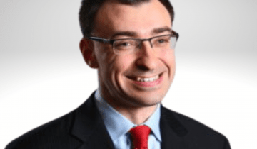Jason has dark brown hair and is smiling wearing a blue shirt, red tie and a blazer.