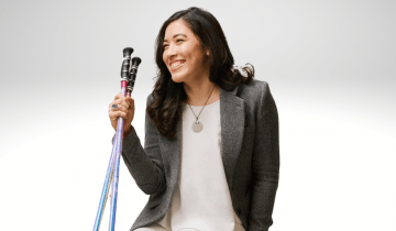 Xian Horn is sitting on a bench smiling, wearing white pants, white shirt and a brown blazer. She has walking sticks in her right hand. She has long brown hair and is smiling.