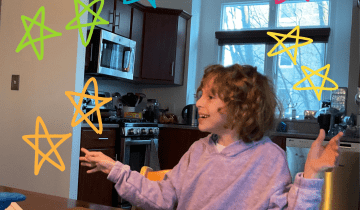 A young girl with medium length curly brown hair is smiling with her arms outstretched while wearing a lilac shirt at a kitchen table in a kitchen of a home.