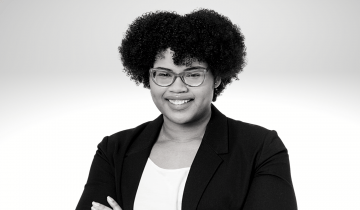 Minelli Vasques has curly brown hair, she is smiling and wearing glasses. She has a white shirt and a black blazer with her arms crossed in front of her.