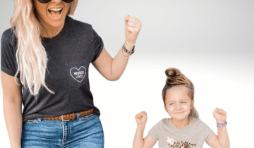 Fifi is a little girl standing in front of her purple walker wearing a grey t-shirt with her hands fisted in the air. She is smiling and has brown hair. She is next to her mom who is wearing a white cowboy hat, dark glasses, has long blonde hair and is smiling wearing a grey shirt and jeans.