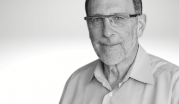 Peter Rosenbaum has dark hair, glasses and a short grey beard. He is wearing a white button down shirt.