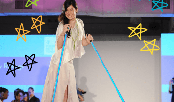 A woman with long brown hair wearing a white flowing dress is on a fashion runway using bright blue poles to walk
