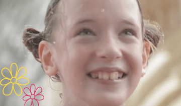 A little girl with blond pigtails and a big smile is in front of a fountain at a water park wearing her swim suit.