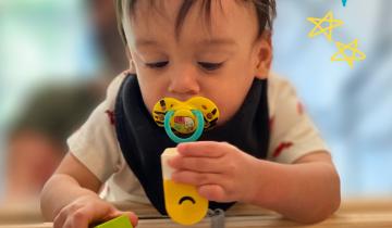 A child with dark hair is sucking on a pacifier while playing with blocks