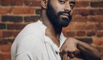 Jerron is standing in front of a brick wall wearing a white shirt with his left hand and arm up in front of his chest.