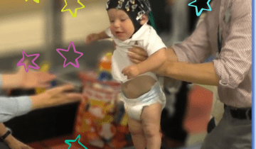 A baby wearing an eeg cap is being held up by a man while he tries to take a step towards a woman with her arms outstretched