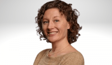 Dr. Heather Riordan has curley shoulder length light brown hair. She is wearing a tan short sleaved shirt and is smiling.
