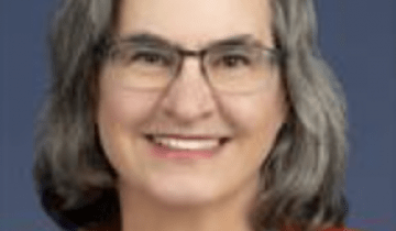 Dr. Kripke has shoulder length salt and pepper hair and is smiling wearing glasses and an orange blazer