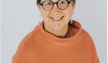 Ginny Paleg headshot. She has short brown hair and is wearing glasses and an orange turtleneck.