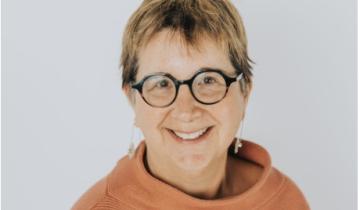 Ginny Paleg headshot. She has short brown hair and is wearing glasses and an orange turtleneck.
