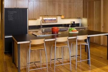 Photo of modern kitchen with wooden paneling. Island bench with 3 stools and black bench top
