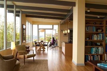 A photo of a modern home with open plan living looking from the lounge room to the dining area and kitchen. There is a young women in a manual wheelchair in the dining room.