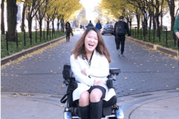 Writer Sarah Kim sitting in her power wheelchair in front of an avenue of trees